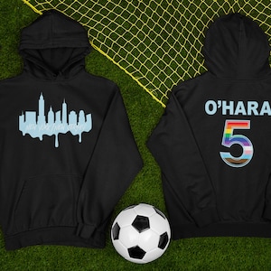 Puede incluir: Dos sudaderas con capucha negras. Una tiene una silueta azul del horizonte de la ciudad de Nueva York con el texto "Nueva York, Nueva Jersey" debajo. La otra sudadera con capucha tiene el texto "O'HARA" y el n&uacute;mero "5" en colores arco&iacute;ris en la parte posterior.