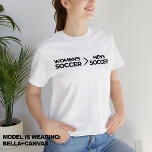 May include: White t-shirt with black text that reads "Women's Soccer > Men's Soccer".