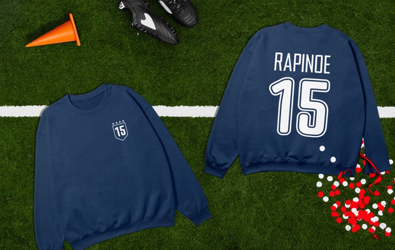 K&ouml;nnte beinhalten: Ein marineblaues Sweatshirt mit dem Namen "RAPINOE" und der Nummer "15" auf der R&uuml;ckseite. Die Vorderseite des Sweatshirts hat einen kleinen wei&szlig;en Schild mit der Nummer "15" darin.