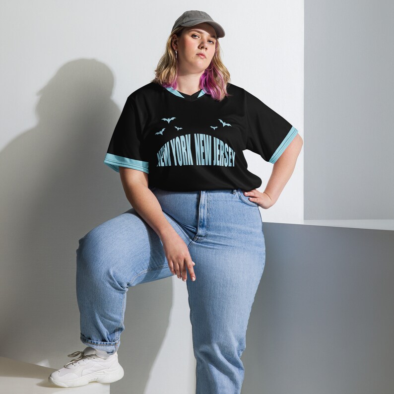 K&ouml;nnte beinhalten: Ein schwarz-blaues Trikot mit dem Text "NEW YORK NEW JERSEY" und ein paar kleinen blauen Flederm&auml;usen, die auf der Vorderseite aufgedruckt sind. Das Model tr&auml;gt eine blaue Jeans und eine schwarz-wei&szlig;e Baseballkappe.