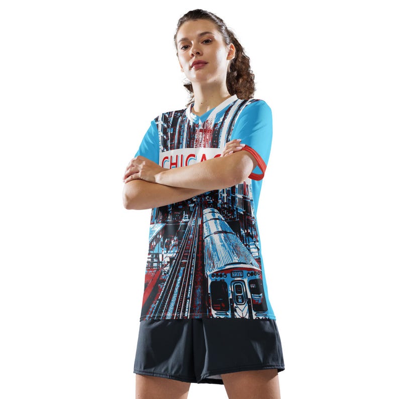 Op de afbeelding: Een blauw en wit shirt met een grafische print van een trein en het woord "CHICAGO" op de voorkant. Het shirt wordt gedragen door een persoon met gekruiste armen.