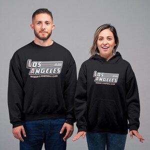 Pu&ograve; includere: Felpa e felpa con cappuccio nere con la scritta "Los Angeles Women's Football Club" in un design stilizzato. Il testo &egrave; in bianco, grigio e rosa. La felpa ha lo scollo tondo e la felpa con cappuccio ha il cappuccio con coulisse.