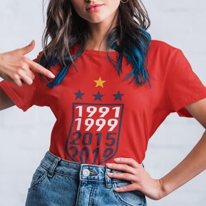 Puede incluir: Camiseta roja con un gr&aacute;fico blanco de un escudo con cuatro estrellas y los a&ntilde;os 1991, 1999, 2015 y 2019.