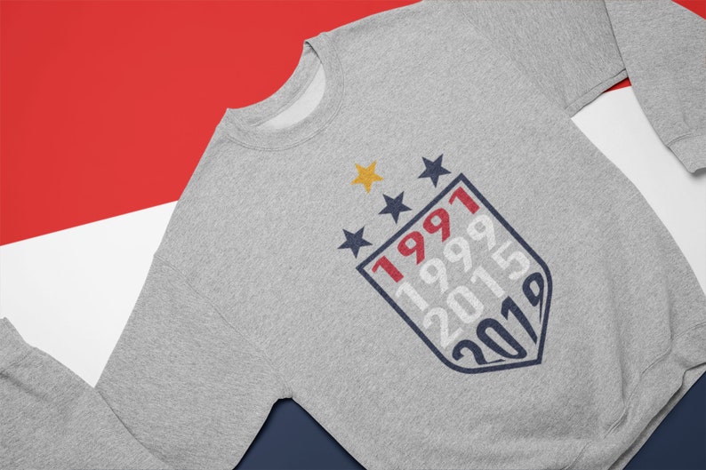 Puede incluir: Sudadera gris con un dise&ntilde;o de escudo azul y rojo con los a&ntilde;os 1991, 1999, 2015 y 2019.