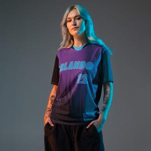 Op de afbeelding: Een vrouw draagt een paars en zwart shirt met de tekst "ORLANDO 22" in blauw. Het shirt heeft een verloopdesign en een zwarte kraag.