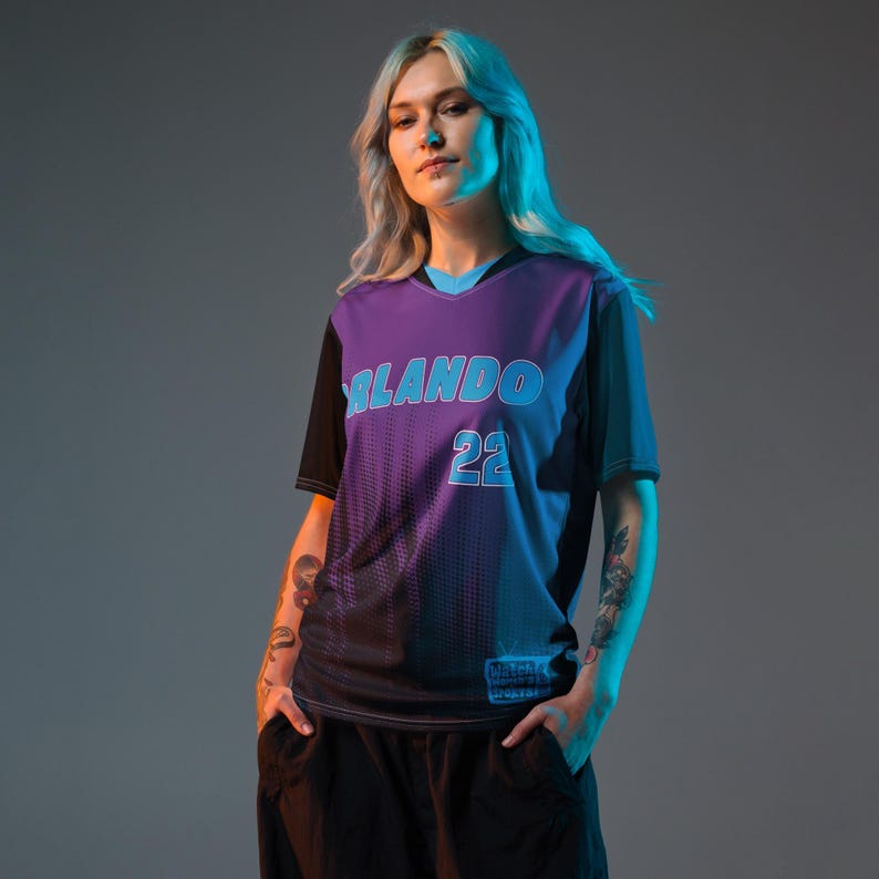 Op de afbeelding: Een vrouw draagt een paars en zwart shirt met de tekst "ORLANDO 22" in blauw. Het shirt heeft een verloopdesign en een zwarte kraag.