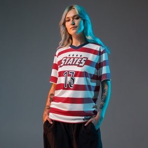 May include: A woman wearing a red and white striped jersey with the word "STATES" and the number "13" on the front. The jersey has a blue collar and blue trim around the sleeves.
