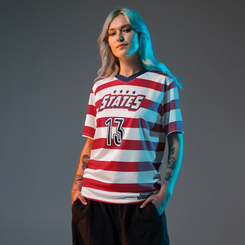 May include: A woman wearing a red and white striped jersey with the word "STATES" and the number "13" on the front. The jersey has a blue collar and blue trim around the sleeves.