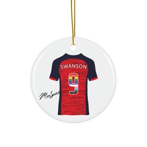 Op de afbeelding: Een ronde, witte keramische ornament met een voetbalshirt ontwerp. Het shirt is rood en zwart met de naam "Swanson" en het nummer "9" in regenboogkleuren. Een gouden draad is bevestigd om op te hangen.