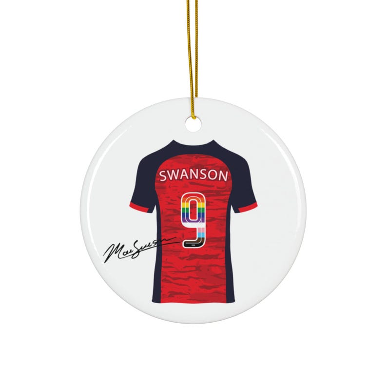 Op de afbeelding: Een ronde, witte keramische ornament met een voetbalshirt ontwerp. Het shirt is rood en zwart met de naam "Swanson" en het nummer "9" in regenboogkleuren. Een gouden draad is bevestigd om op te hangen.