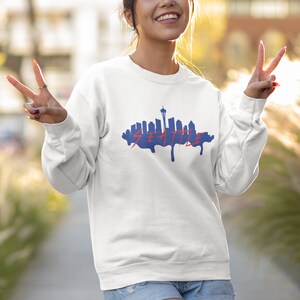 May include: A white sweatshirt with a blue silhouette of the Seattle skyline and the word "SEATTLE" in red dripping paint. The model is wearing the sweatshirt and smiling.