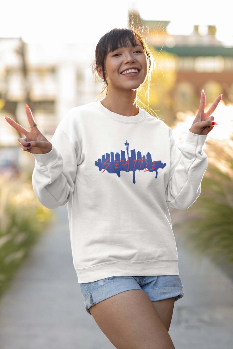 May include: A white sweatshirt with a blue silhouette of the Seattle skyline and the word "SEATTLE" in red dripping paint. The model is wearing the sweatshirt and smiling.