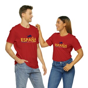 May include: Two people wearing red t-shirts with the word "ESPA&Ntilde;A" and a bull graphic printed on the front. The shirts are a bright red color and the text is in a yellow and blue font.
