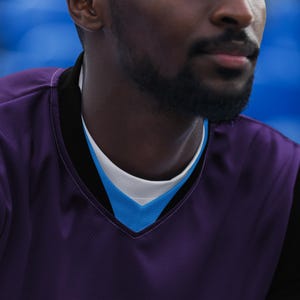 Op de afbeelding: Een close-up van een persoon die een paars shirt draagt met een blauwe en witte kraag. Het shirt heeft een v-hals en een textuur.