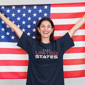 Op de afbeelding: Een persoon draagt een donkerblauw T-shirt met de tekst "UNITED STATES" in rood en wit. Ze houdt een grote Amerikaanse vlag vast.