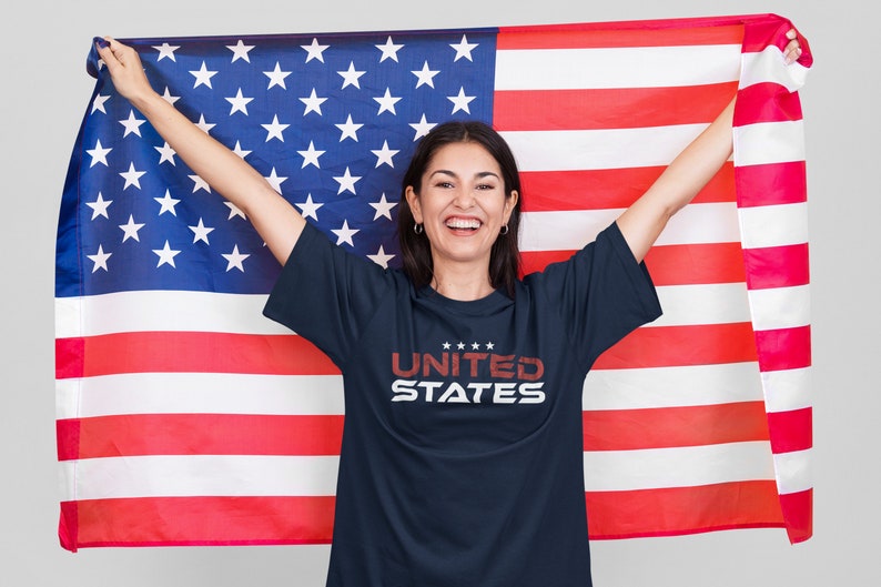 Op de afbeelding: Een persoon draagt een donkerblauw T-shirt met de tekst "UNITED STATES" in rood en wit. Ze houdt een grote Amerikaanse vlag vast.