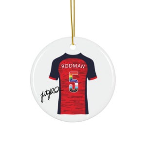 Op de afbeelding: Een ronde, witte keramische kerstversiering met een rood-zwart voetbalshirtontwerp. Het shirt heeft de naam "RODMAN" boven een regenboogkleurig nummer "5". Er is een gouden touwtje bevestigd om op te hangen. De versiering heeft een zwarte handtekening.