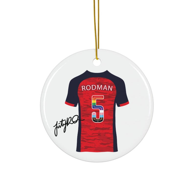 Op de afbeelding: Een ronde, witte keramische kerstversiering met een rood-zwart voetbalshirtontwerp. Het shirt heeft de naam "RODMAN" boven een regenboogkleurig nummer "5". Er is een gouden touwtje bevestigd om op te hangen. De versiering heeft een zwarte handtekening.