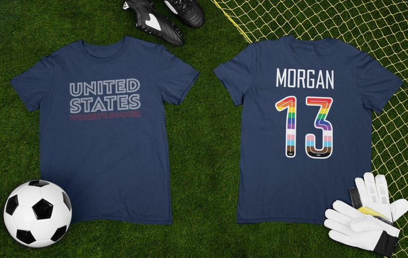 K&ouml;nnte beinhalten: Zwei marineblaue T-Shirts mit dem Text "UNITED STATES WOMEN'S SOCCER" auf der Vorderseite und der Nummer "13" mit dem Namen "MORGAN" auf der R&uuml;ckseite. Die Nummer "13" ist in Regenbogenfarben umrandet.