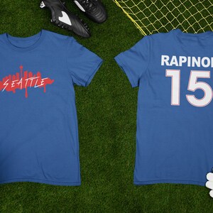 Puede incluir: Dos camisetas azules, una con un gr&aacute;fico rojo del horizonte de Seattle y la otra con el nombre "RAPINOE" y el n&uacute;mero "15" en blanco. Un bal&oacute;n de f&uacute;tbol y botas de f&uacute;tbol est&aacute;n sobre la hierba verde.