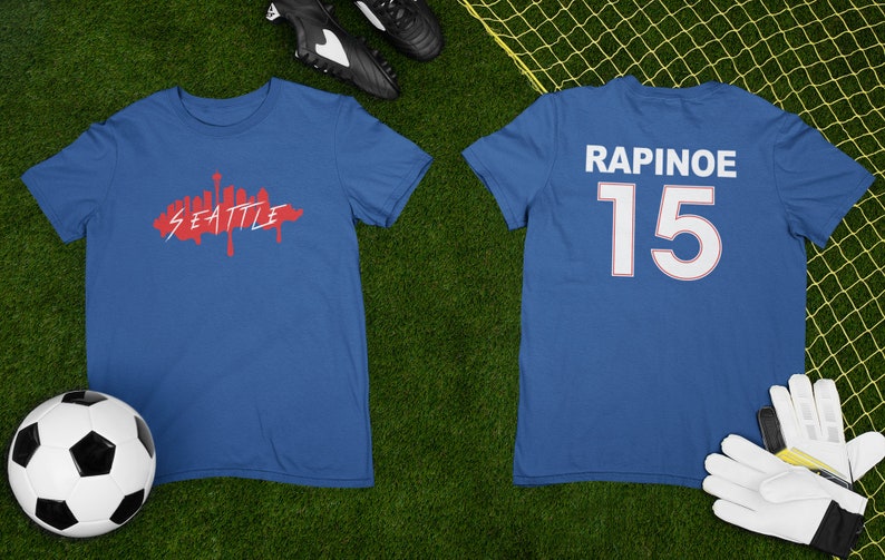 Puede incluir: Dos camisetas azules, una con un gr&aacute;fico rojo del horizonte de Seattle y la otra con el nombre "RAPINOE" y el n&uacute;mero "15" en blanco. Un bal&oacute;n de f&uacute;tbol y botas de f&uacute;tbol est&aacute;n sobre la hierba verde.