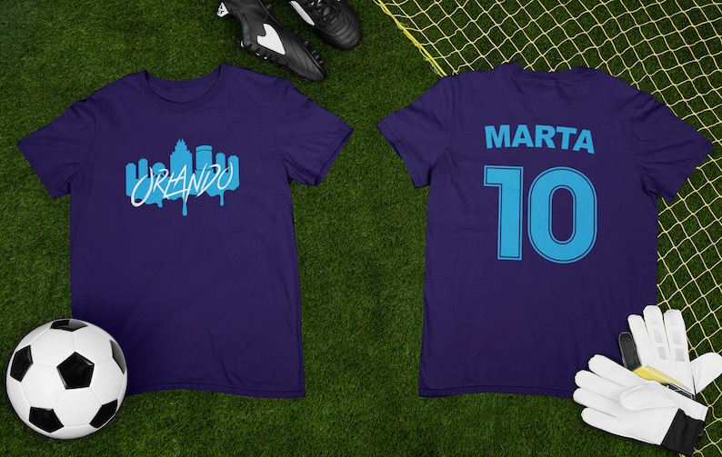 May include: A purple soccer jersey with a blue skyline graphic and the word "ORLANDO" on the front. The back of the jersey has the name "MARTA" and the number "10" in blue.