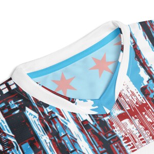 Op de afbeelding: Wit T-shirt met een blauw en rood stadsbeeldontwerp. De kraag is wit met een lichtblauwe binnenvoering. Het ontwerp heeft rode sterren op de binnenvoering.