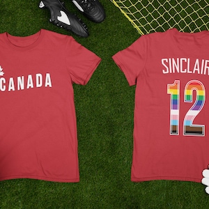May include: Two red t-shirts with white text and a football on a green field. One shirt says "CANADA" with a maple leaf and the number 12. The other shirt says "SINCLAIR 12" with a rainbow and progress pride flag.