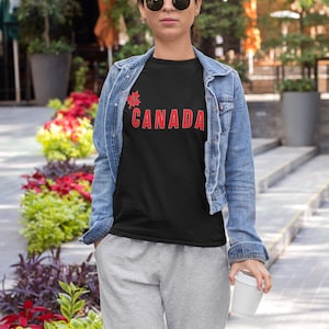 May include: A black t-shirt with the word "CANADA" in red, and a red maple leaf graphic. The person is wearing a denim jacket, grey sweatpants, and sunglasses. They are holding a white coffee cup.