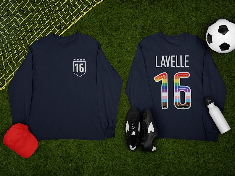 Op de afbeelding: Twee marineblauwe shirts met lange mouwen met het nummer 16 erop. E&eacute;n shirt heeft het nummer in een wit schild met sterren erboven. Het andere shirt heeft het nummer in regenboogkleuren met de naam "Lavelle" erboven.