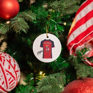 Op de afbeelding: Een ronde, witte kerstversiering met een rood en blauw voetbalshirt met de naam "RODMAN" en het nummer "5". De versiering hangt aan een kerstboom met rode en witte decoraties.