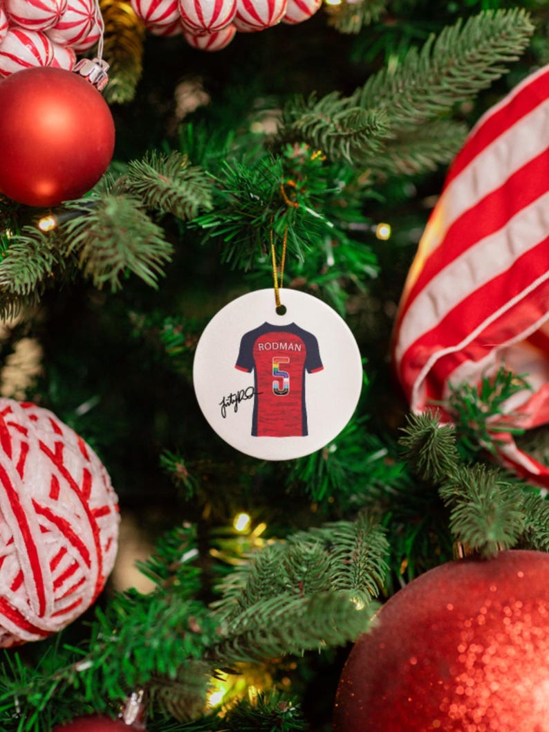 Op de afbeelding: Een ronde, witte kerstversiering met een rood en blauw voetbalshirt met de naam "RODMAN" en het nummer "5". De versiering hangt aan een kerstboom met rode en witte decoraties.