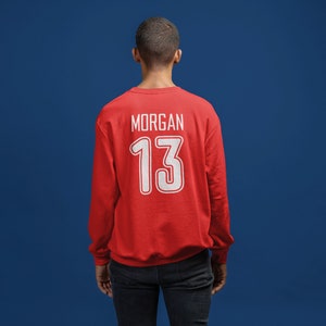 K&ouml;nnte beinhalten: Roter Pullover mit wei&szlig;em Text auf der R&uuml;ckseite, der "MORGAN 13" lautet.