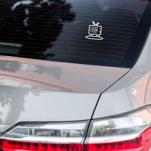 May include: A white car with a sticker on the back window. The sticker is a black and white illustration of a television with the text "Watch Woman's Sports" inside.