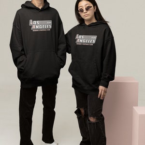 Pu&ograve; includere: Due persone indossano felpe con cappuccio nere con la scritta "Los Angeles Women's Football Club" in argento e rosso. Indossano anche pantaloni neri e scarpe da ginnastica bianche. Una persona indossa un cappello da pescatore rosa.