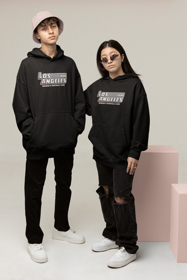 Pu&ograve; includere: Due persone indossano felpe con cappuccio nere con la scritta "Los Angeles Women's Football Club" in argento e rosso. Indossano anche pantaloni neri e scarpe da ginnastica bianche. Una persona indossa un cappello da pescatore rosa.