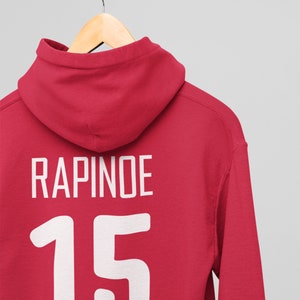 Może przedstawiać: Czerwona bluza z kapturem z nadrukiem "RAPINOE" i numerem "15" na plecach w kolorze białym.