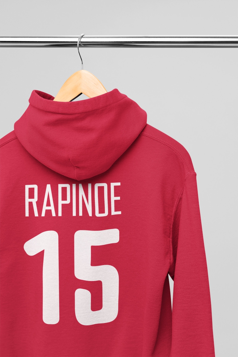 Może przedstawiać: Czerwona bluza z kapturem z nadrukiem "RAPINOE" i numerem "15" na plecach w kolorze białym.
