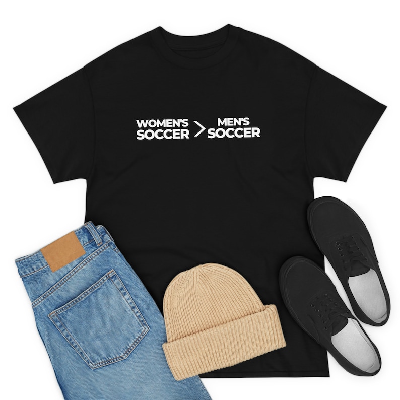 May include: Black t-shirt with white text that reads "Women's Soccer > Men's Soccer". The t-shirt is displayed with blue jeans, a beige beanie, and black shoes.