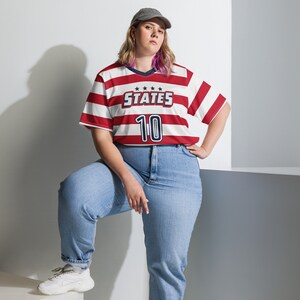 May include: A woman wearing a red and white striped jersey with the text "STATES 10" on the front. She is wearing blue jeans and a baseball cap.
