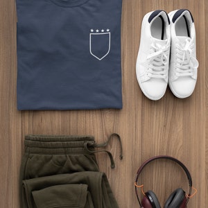 May include: A navy blue t-shirt with a white pocket featuring five stars on a shield. A pair of white sneakers, olive green sweatpants, and a pair of burgundy headphones are also pictured.