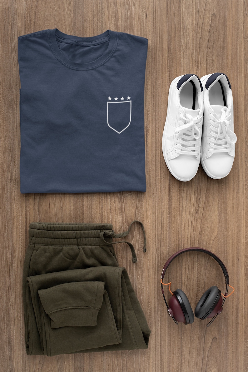 May include: A navy blue t-shirt with a white pocket featuring five stars on a shield. A pair of white sneakers, olive green sweatpants, and a pair of burgundy headphones are also pictured.