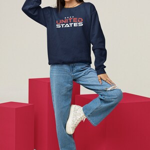 Op de afbeelding: Een donkerblauw sweatshirt met de tekst "UNITED STATES" in witte en rode letters. Het model draagt een blauwe jeans en witte sneakers.