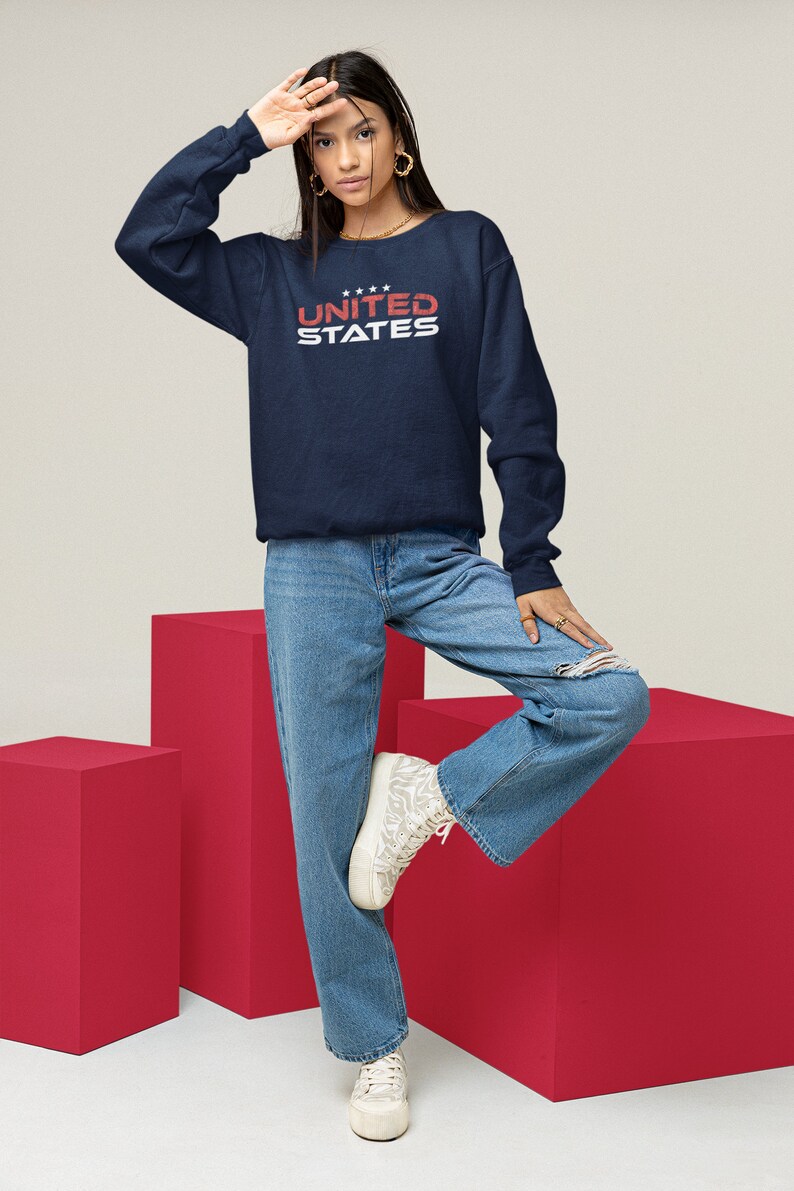 Op de afbeelding: Een donkerblauw sweatshirt met de tekst "UNITED STATES" in witte en rode letters. Het model draagt een blauwe jeans en witte sneakers.