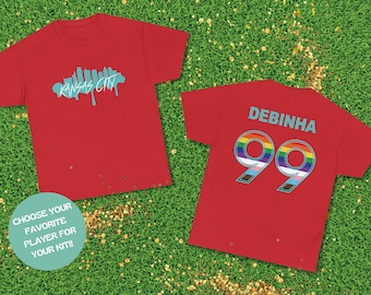 May include: Two red t-shirts, one with a blue graphic of the Kansas City skyline and the text "KANSAS CITY", and the other with the text "DEBÍNHÁ" and the number "99" with a rainbow design.