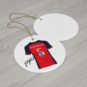 Op de afbeelding: Twee witte keramische schijfversieringen met bruine touwtjes. E&eacute;n heeft een rood en marineblauw jersey-ontwerp met de naam "RODMAN" en het nummer "5" in regenboogkleuren. De andere versiering is effen wit.