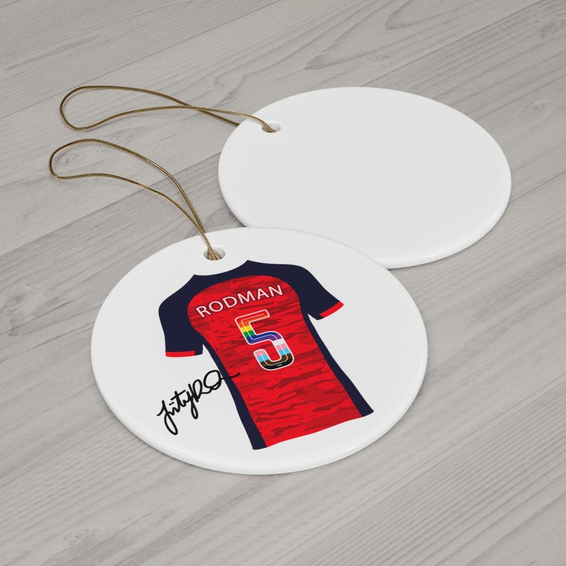 Op de afbeelding: Twee witte keramische schijfversieringen met bruine touwtjes. E&eacute;n heeft een rood en marineblauw jersey-ontwerp met de naam "RODMAN" en het nummer "5" in regenboogkleuren. De andere versiering is effen wit.