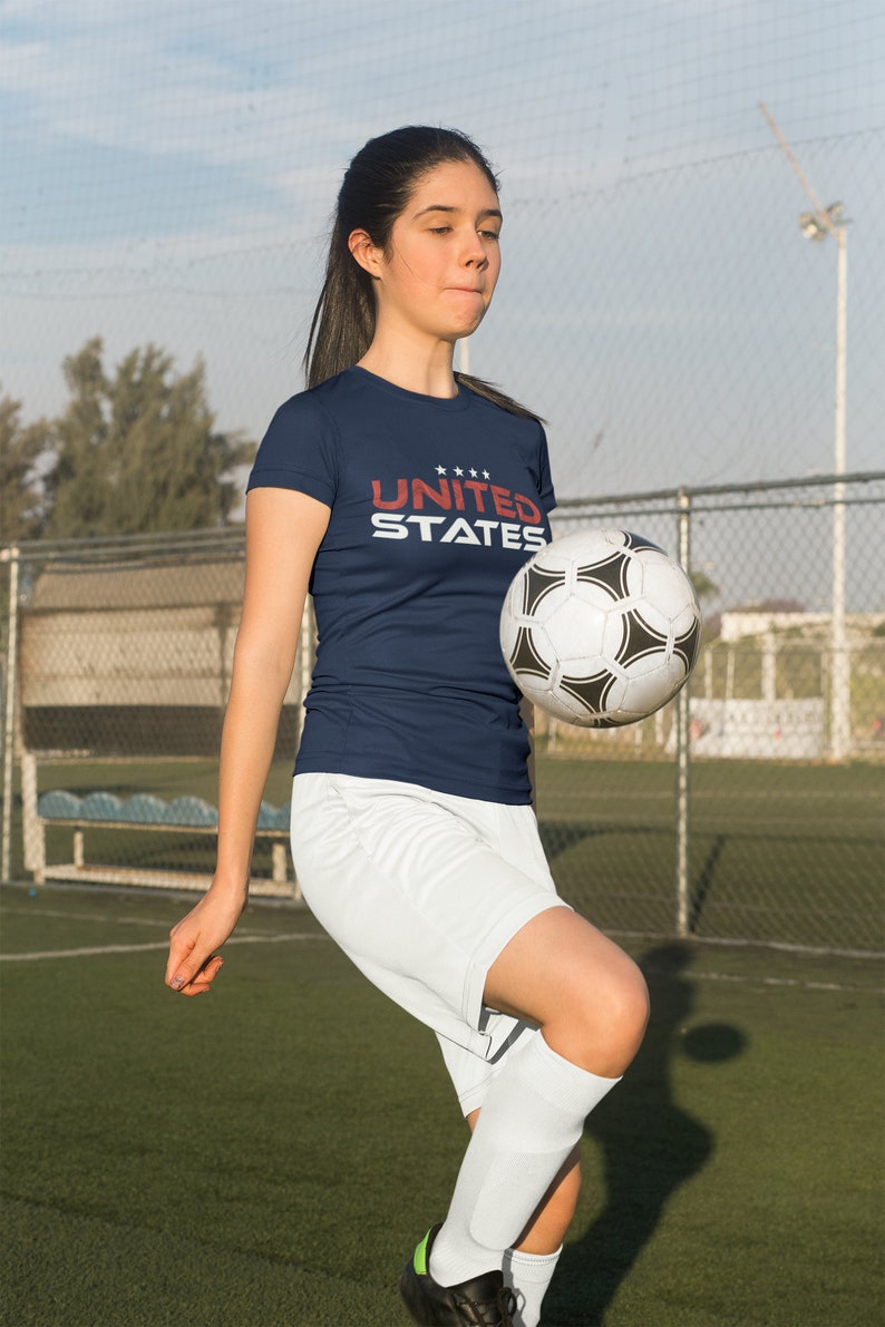 Op de afbeelding: Een vrouw draagt een donkerblauw T-shirt met de tekst "UNITED STATES" in rood en wit. Ze houdt een witte en zwarte voetbal vast en draagt een witte broek en sokken.