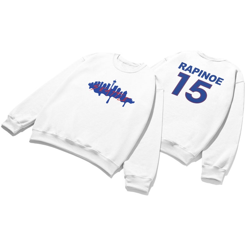 May include: White sweatshirt with a blue and red Seattle skyline graphic on the front and the text "RAPINOE 15" in blue on the back.