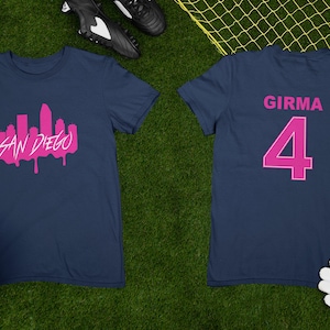 May include: Two navy blue t-shirts with pink lettering. The front of one shirt has a pink silhouette of the San Diego skyline with the words "SAN DIEGO" underneath. The back of the other shirt has the name "GIRMA" and the number "4" in pink.